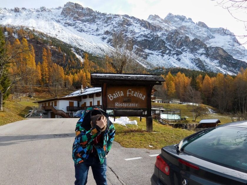 Dónde alojarse en los Dolomitas, Italia: mejores zonas y hoteles baita fraina where to stay in the dolomites italy Cartel de Baita Fraina en Cortina d'Ampezzo, con un amigo inclinado junto a él y una montaña nevada al fondo, Dolomitas, Italia